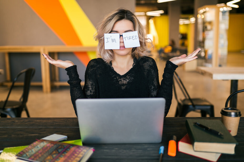 portrait of young pretty tired woman with paper stickers on glasses sitting at table in black shirt working on laptop in co-working office, funny face expression, frustrated emotion personalistka